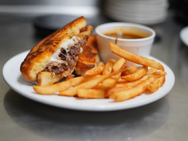 A birria grilled cheese cut in half, served with french fries and a cup of consomé.