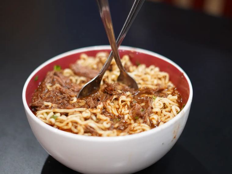 A bowl of birria ramen with shredded beef, noodles and two spoons.