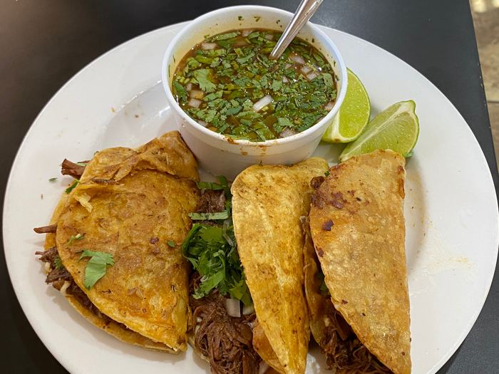 Three quesabirria tacos on a plate with a cup of consommé and lime wedges.