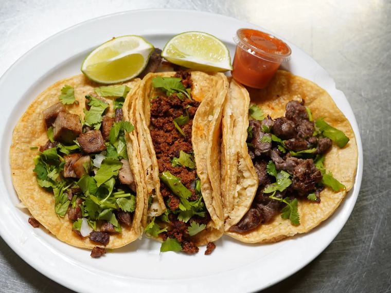 Three Mexi style tacos on corn tortillas with cilantro, onion and lime wedges.